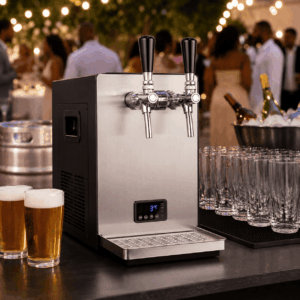 Une tireuse à bières PRO à deux becs (comme dans un vrai bar à bières), en acier inoxydable, trône sur le comptoir, entourée de verres et de deux bières déjà servies. Les convives discutent dans une ambiance feutrée, qui rappelle un événement ou une fête en plein air.