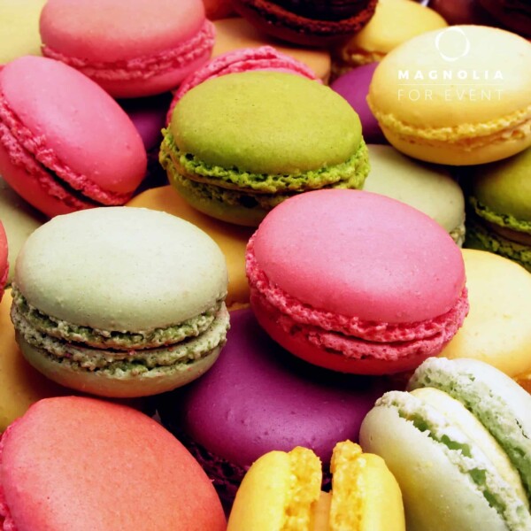 Un gros plan de notre PLATEAU DE 48 PETITS FOURS SUCRÉS DÉLICATS montre des macarons roses, verts, jaunes, orange et violets empilés, avec un subtil logo "Magnolia For Event" dans le coin supérieur droit.