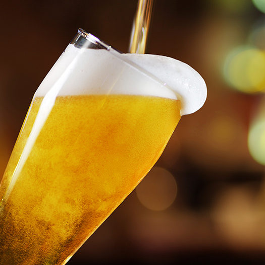 Gros plan sur une bière dorée s'écoulant de la TIREUSE À BIÈRES PRO 2 BECS (Comme dans un vrai bar à bières) dans un verre incliné, formant une mousse épaisse, avec des lumières floues et chaudes en arrière-plan.