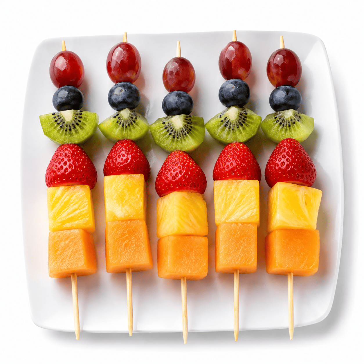 Vingt BROCHETTES DE FRUITS, composées chacune de morceaux d'ananas, de melon, de fraise, de kiwi, de myrtille et de raisin, sont disposées avec art sur une assiette carrée blanche, sur un fond blanc.