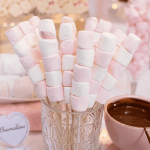 BROCHETTE DE GUIMAUVE — des guimauves roses et blanches enfilées sur des brochettes et présentées dans un verre, accompagnées d'un bol de chocolat fondu. Des décorations aux couleurs pastel et une étiquette floue apparaissent en arrière-plan.