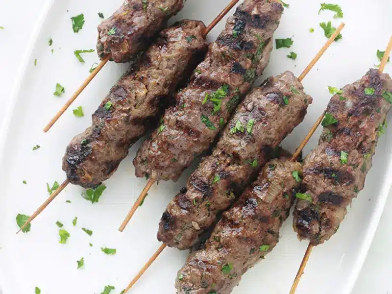 Six brochettes BROCHETTE VEGETARIENNE BARBECUE, idéales pour les événements barbecue, sont disposées sur une assiette blanche, avec des marques de grill visibles et une texture savoureuse et dorée.