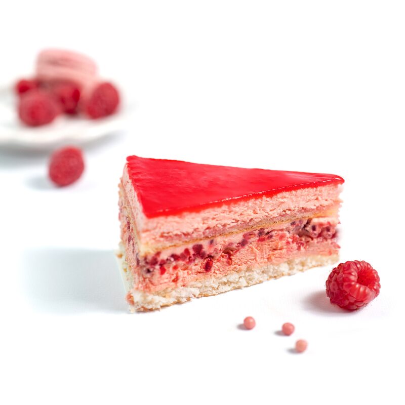Un gâteau FRAMBOISINE 12/16 PARTS à couches de framboises avec un glaçage rose brillant est mis en évidence, entouré de framboises entières et de petits bonbons roses sur une surface blanche. À l'arrière-plan, une assiette contenant d'autres framboises et un macaron est floue.