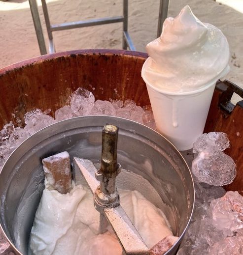 Un seau en bois avec de la glace contient une baratte en métal contenant de la glace à la vanille ANIMATION SORBET COCO ANTILLAIS (1h30). Un gobelet de soft serve est posé sur la glace à côté du baratteur, avec du sable visible en arrière-plan.