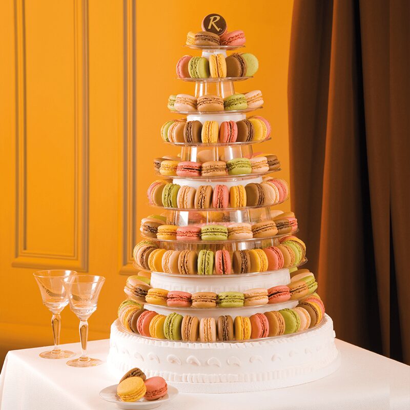 La PIÉCE MONTÉE PYRAMIDE DE 91 MACARONS est une haute tour de macarons avec des macarons colorés empilés en étages sur un gâteau blanc, placée sur une nappe blanche avec deux verres à vin vides et des macarons supplémentaires près d'un mur orange.