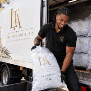 Un homme souriant, portant des gants noirs, décharge un SAC DE GLAÇONS DE 20 KG (pour remplir la cuve et refroidir les bouteilles) d'un camion portant l'enseigne « Le Traiteur Antillais », orné d'un logo doré et de motifs de feuilles tropicales. D'autres sacs de glaçons sont empilés à l'intérieur du camion.
