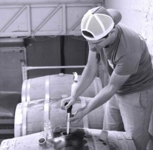 Une personne portant une casquette de baseball à l'envers utilise un outil pour extraire le FÛT DE BIÈRE BLONDE 5° 30L (Bière artisanale bouledogue) d'un tonneau en bois dans un environnement industriel, avec plusieurs tonneaux et petits récipients en verre visibles.