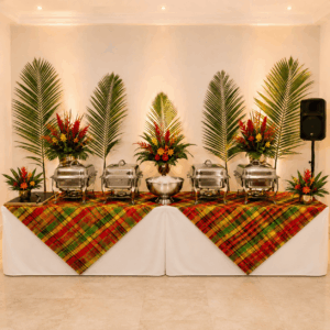 UN BUFFET À L'AMBIANCE ANTILLAISE, agrémenté de quatre réchauds, d'un grand saladier en métal, de compositions florales tropicales et de feuilles de palmier. La table est recouverte d'une nappe blanche et de napperons à carreaux aux couleurs vives.