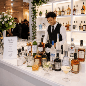 Un barman souriant se tient derrière un comptoir bien éclairé où sont exposées diverses bouteilles d'alcool, un cocktail et des verres. Une pancarte posée sur le comptoir indique « CHOISISSEZ LES HARDS POUR VOTRE ÉVÉNEMENT », présentant différentes options de boissons tandis que des personnes discutent en arrière-plan.