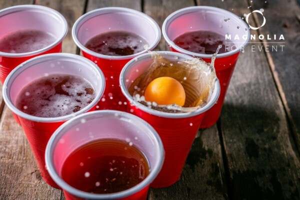 Une balle de ping-pong jaune éclabousse un gobelet de bière rouge au milieu d'autres gobelets sur une table en bois, mettant en évidence le jeu BEER PONG en action.