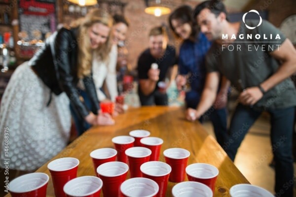 Un groupe de jeunes adultes joue au BEER PONG, en visant avec une balle de ping-pong un triangle de gobelets rouges sur une table en bois, dans une atmosphère animée et amusante.
