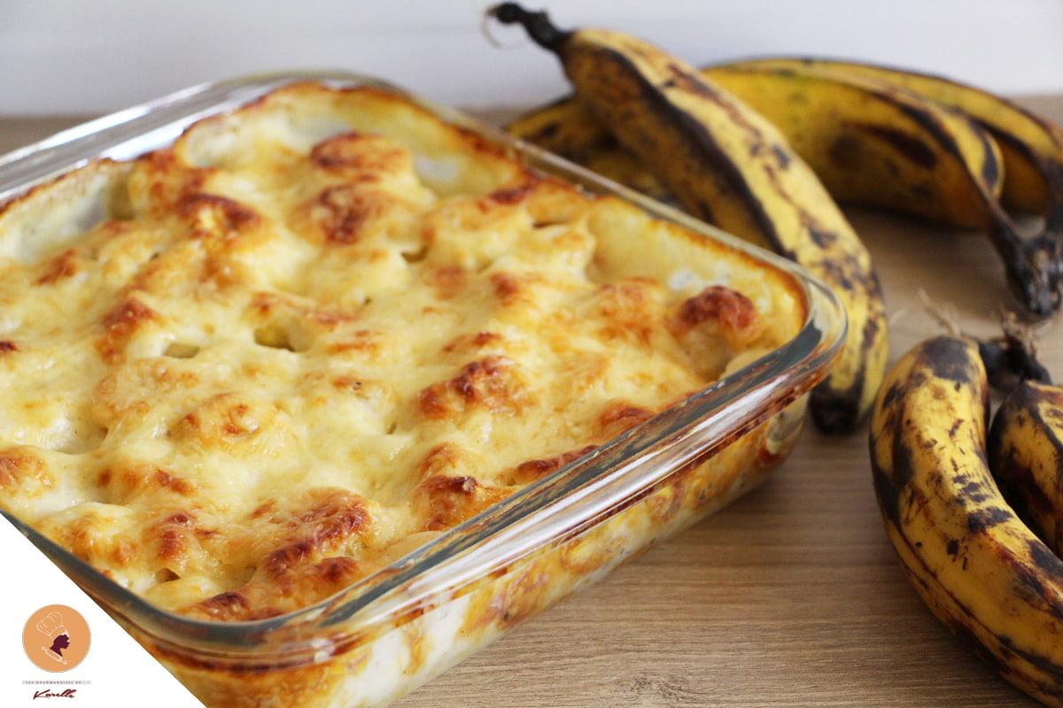 Une portion de GRATIN DE BANANES PLANTIN (250 g la part) est posée dans un plat à four en verre, dorée et fromagée, sur une surface en bois, à côté de plusieurs bananes plantains mûres.