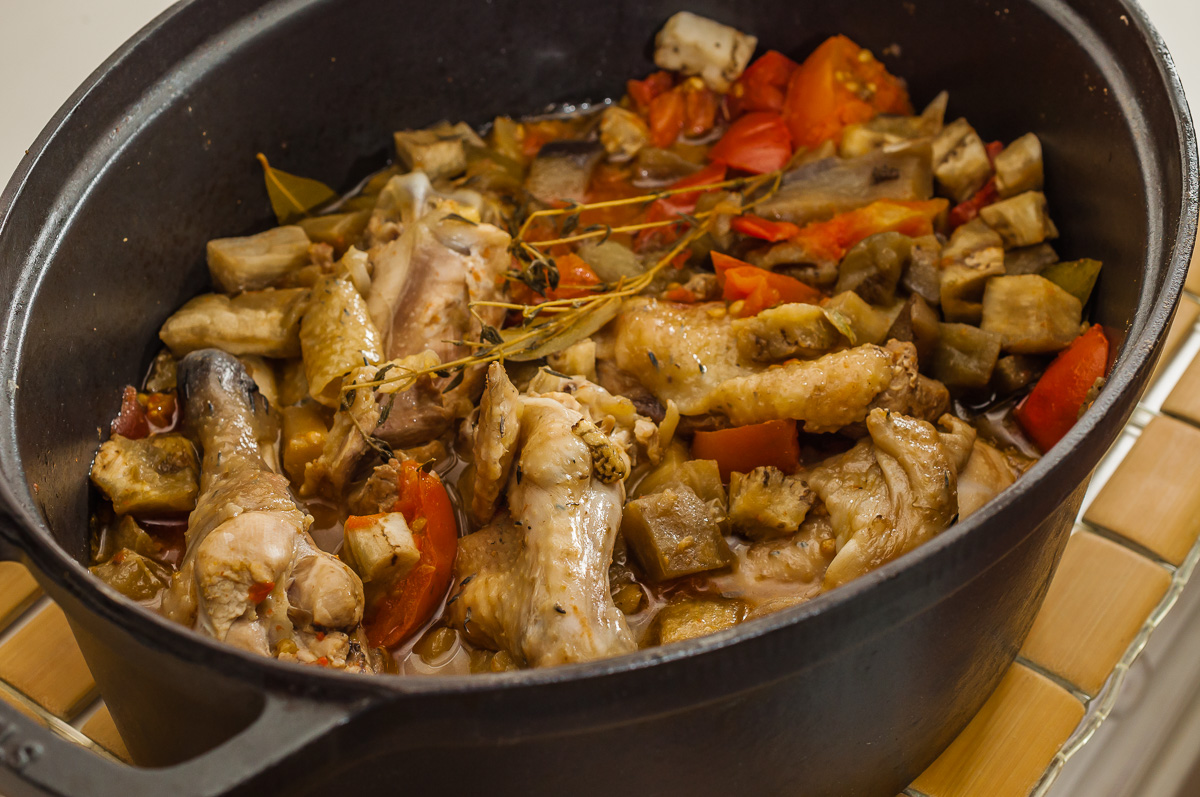 Un pot noir rempli de PASTEL DE VIANDE (Barquette de 10 pièces), de tomates en dés, d'aubergines et d'herbes est posé sur une natte en bambou.