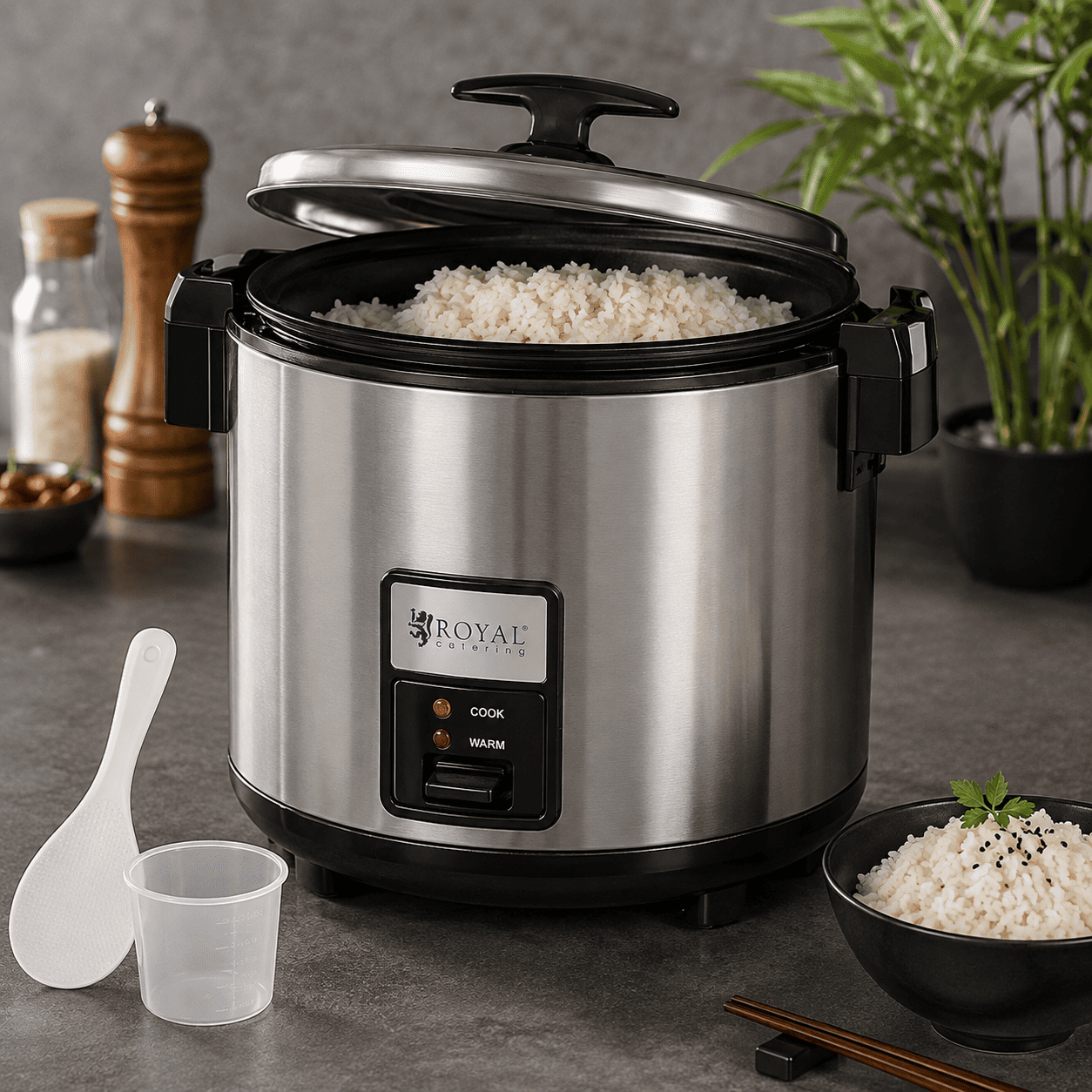 Une MACHINE À RIZ remplie de riz cuit à la perfection trône sur le plan de travail de la cuisine. À proximité se trouvent une spatule à riz, un verre doseur, un bol de riz, des baguettes et des ingrédients tels que du sel et du poivre.