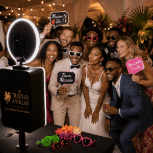 Une joyeuse fête de mariage se rassemble autour du PHOTOBOOTH FIXE (Pour des souvenirs inoubliables à ramener à la maison), prenant des poses enjouées avec des pancartes et des accessoires ludiques. Le groupe, vêtu d'élégantes tenues de cérémonie, sourit à l'objectif devant un décor tropical et des accessoires aux couleurs vives disposés sur la table devant eux.