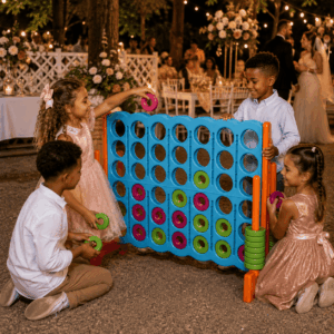 Quatre enfants, sur leur trente-et-un pour une occasion spéciale, jouent au PUISSANCE 4 GÉANT dans un décor festif en plein air, agrémenté de fleurs et de lumières, avec des invités en arrière-plan. Souriant et s'amusant avec les pièces colorées du jeu, les enfants s'amusent ensemble avec cette version géante du Connect Four.
