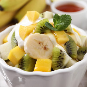 SALADE DE FRUITS EXOTIQUES : Salade de fruits frais composée de tranches de bananes, de kiwis, de mangues et de pommes, garnie de yaourt et d'une feuille de menthe. Des bananes et un petit plat en sauce apparaissent en arrière-plan.
