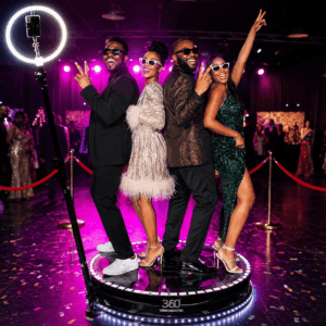 Quatre personnes vêtues de tenues élégantes prennent la pose et sourient sur la plateforme VIDÉOBOOTH À 360°, lors d'un événement animé, avec une lampe annulaire et un éclairage violet en arrière-plan.