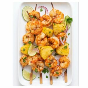 BROCHETTES ANTILLAISES À L'ANANAS ET AUX CREVETTES, agrémentées de coriandre fraîche, d'oignon rouge émincé et de quartiers de citron vert, servies sur une assiette rectangulaire blanche.