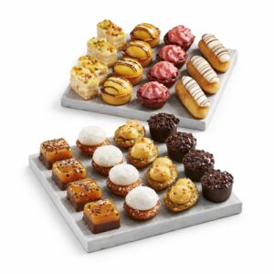 Le PLATEAU DE 48 PETITS FOURS ART DÉCO présente deux plateaux de pâtisseries assorties - mini éclairs, tartelettes aux fruits, carrés au caramel, gâteaux au chocolat et pâtisseries glacées - joliment disposés sur des planches gris clair.