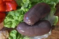 Deux saucisses BOUDIN ANTILLAIS 80 GR (commande minimum de 5 pièces) sont placées sur des feuilles de laitue verte, avec des morceaux de poivron rouge et une petite quantité de condiment léger visible à proximité sur une surface en bois.