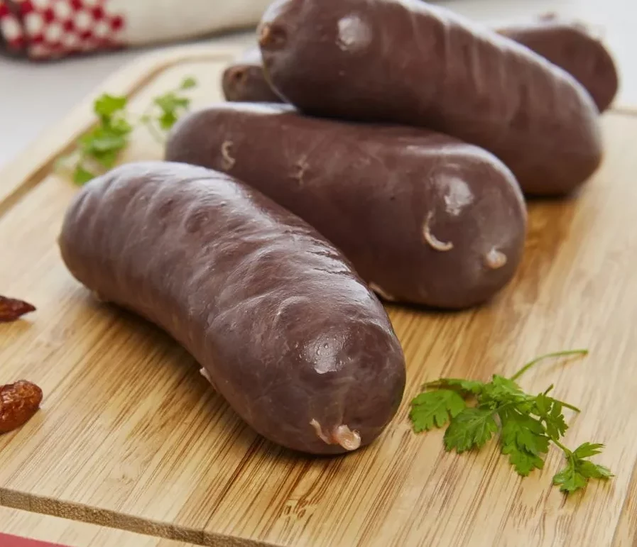 Trois saucisses BOUDIN ANTILLAIS 80 GR, foncées et dodues (commande minimum de 5 pièces), sont disposées sur une planche à découper en bois, à côté d'herbes fraîches, avec un tissu rouge et blanc flou en arrière-plan.