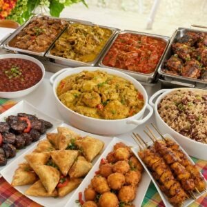 Un « buffet antillais “CARAÏBES” : entrée, plat » pour un minimum de 10 personnes, proposant un large choix de spécialités des Caraïbes telles que le riz aux pois, des ragoûts, du poulet grillé, des samosas, des bananes plantains frites, des brochettes, des boulettes et des haricots — le tout joliment présenté sur des assiettes blanches posées sur une nappe colorée.