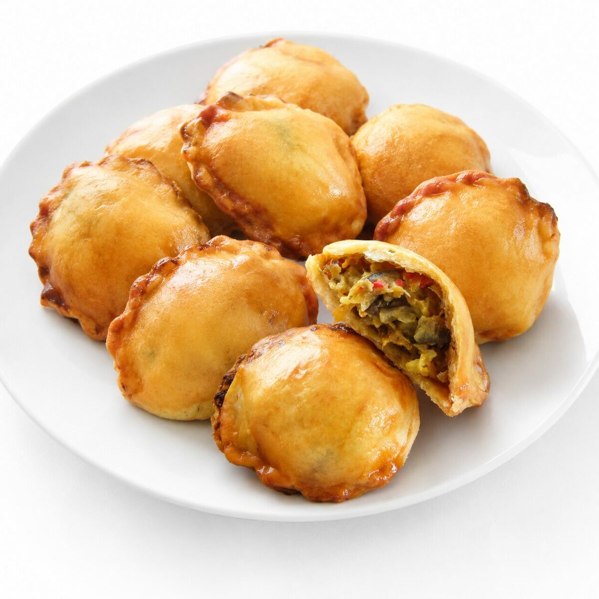 Une assiette blanche présente sept MINI PÂTÉ ANTILLAIS AU BOEUF d'un brun doré, dont l'un est entaillé, dévoilant une savoureuse farce au bœuf.