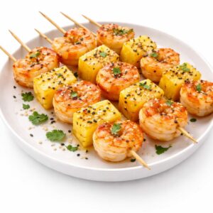 ANANAS VICTORIA + BROCHETTE DE CREVETTES (300g) : Brochettes de crevettes et d'ananas grillés, garnies de graines de sésame et de coriandre fraîche, joliment disposées sur une assiette blanche.