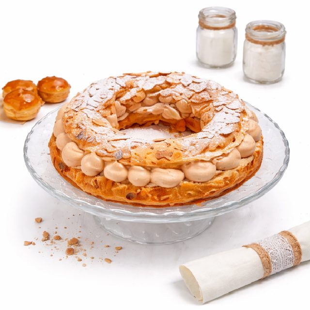 Le PARIS BREST (10 pièces), un cercle pâtissier garni d'amandes tranchées et de sucre en poudre et fourré de praliné crémeux, est présenté sur un présentoir à gâteaux en verre. Deux bocaux, des petites pâtisseries et une serviette roulée apparaissent en arrière-plan.