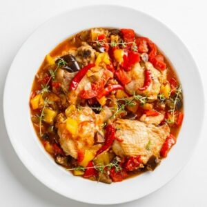 Une assiette blanche met en valeur le KEDJÉNOU DE POULET (300 g), composé de poulet tendre, de poivrons rouges et jaunes, de tomates et d'herbes aromatiques mijotés dans une sauce onctueuse, le tout sur un fond blanc immaculé.