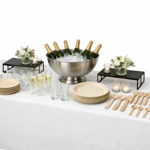 Une table dressée pour une fête, avec le GRAND PACK TRAITEUR pour une organisation parfaite du buffet : seau à champagne, piles d'assiettes, verres, couverts en bois, petits bouquets de fleurs et bougies votives, le tout sur une nappe blanche.
