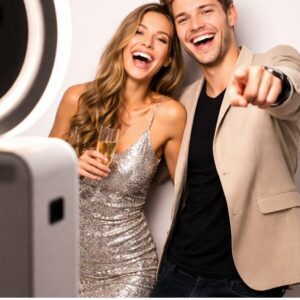 Deux personnes sourient et prennent la pose pour une photo prise dans un PHOTOBOOTH FIXE (Pour des souvenirs inoubliables à ramener à la maison). La femme, vêtue d'une robe argentée pailletée, tient un verre à la main, tandis que l'homme, vêtu d'un blazer beige, pointe l'appareil photo d'un air espiègle. Tous deux ont l'air heureux et festifs alors qu'ils créent des souvenirs inoubliables.