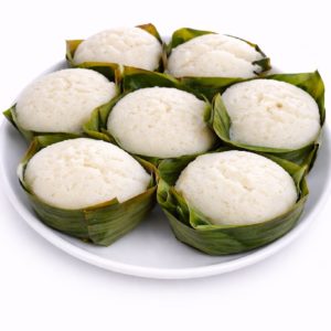 Une assiette blanche présente ABLO (200 g/pers) - sept gâteaux de riz cuits à la vapeur, ronds et moelleux, présentés dans des feuilles de bananier, disposés en rangs serrés et présentant une texture légère et moelleuse.