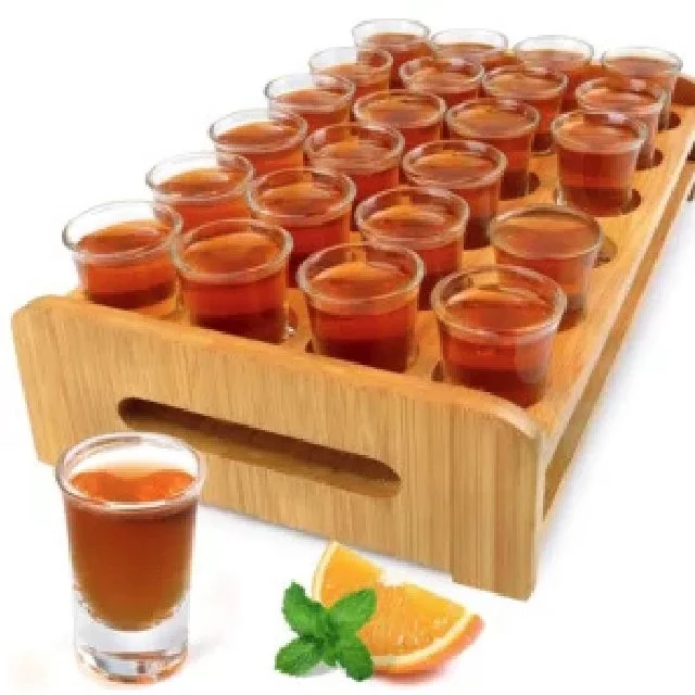 Le PLATEAU DE 24 VERRES À TI PUNCH est un plateau en bois qui contient 24 verres à liqueur remplis en rangées, avec un verre à liqueur devant à côté d'un quartier d'orange et de feuilles de menthe.