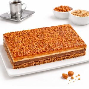 Le CROQUANT AU CARAMEL (12/16 parts), un dessert à couches rectangulaires avec une garniture de noix caramélisées, est placé sur un plateau blanc. Une tasse de café et deux bols de noix entières et hachées sont visibles à l'arrière-plan.