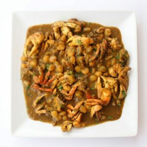 Une assiette carrée blanche présente un « DOMBRÉ DE CRABES EN COLOMBO » — un ragoût brun copieux composé de morceaux de crabe, de pois chiches et d'herbes aromatiques —, servi sur un fond blanc uni.