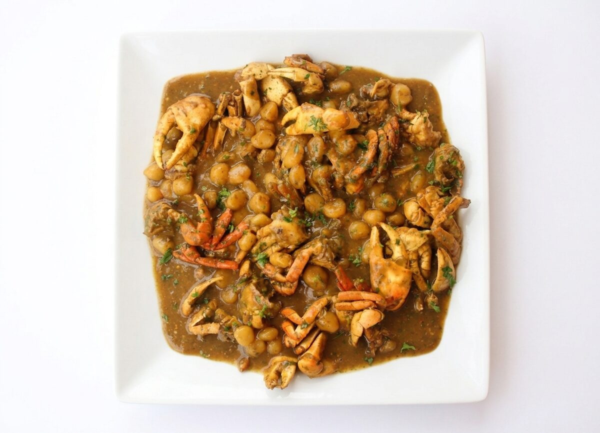 Une assiette carrée blanche présente un « DOMBRÉ DE CRABES EN COLOMBO » — un ragoût brun copieux composé de morceaux de crabe, de pois chiches et d'herbes aromatiques —, servi sur un fond blanc uni.