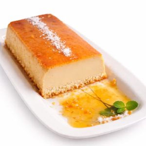 Le FLAN COCO FAIT MAISON (Gâteau de 10 parts) est un dessert rectangulaire au flan de coco avec un glaçage au caramel, servi sur une assiette blanche et décoré de flocons de noix de coco, de sirop et d'un brin de menthe.