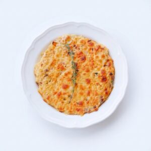 Un plat rond de GRATIN D’IGNAMES (commande minimum : 5 portions – 250 g par portion) recouvert d’une croûte dorée et fondante, agrémenté d’un brin d’herbes aromatiques, servi sur une assiette blanche devant un fond blanc.