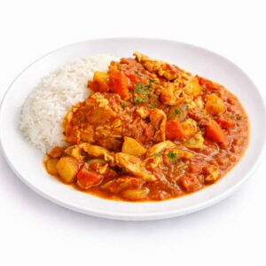Une portion de MAFÉ (300 g) dans une assiette blanche comprend du riz blanc cuit à la vapeur avec du poulet, des pommes de terre et des carottes dans une riche sauce rouge-orange, agrémentée d'herbes vertes.