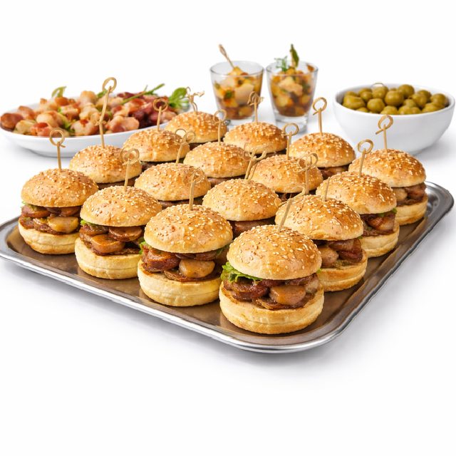 Un plateau métallique présente des MINI-BURGERS AU FOIE GRAS (commande minimum de 20 pièces, 25g/pc), chaque burger étant accompagné d'un petit pain aux graines de sésame et d'une brochette. À l'arrière-plan, des bols d'olives, des amuse-gueule et des desserts en couches.