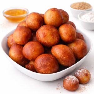 Un bol blanc rempli de PUFF PUFF (Minimum de commande 10 pièces) est posé sur une surface blanche, entouré de petits bols de miel, de farine et de sucre. Certains PUFF PUFF sont saupoudrés de sucre en poudre.
