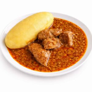 Une assiette blanche avec du FOUTOU (200g/pers), un aliment jaune ressemblant à de la pâte, et de la soupe egusi comprenant des morceaux de viande dans une sauce épaisse à base de graines de melon orange.
